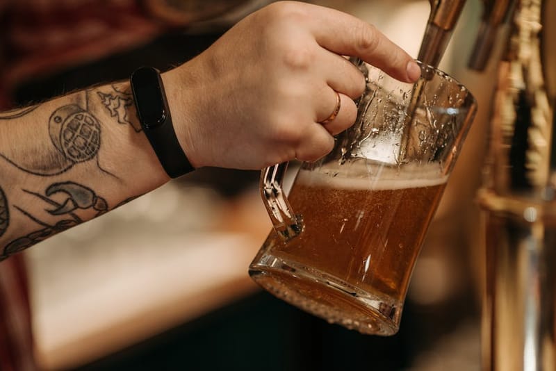 Close-up of a tattooed hand pouring beer into a glass mug from a draft tap.
